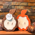Three fall-themed decorative candles displayed on a wooden shelf against a rustic stone wall. The set includes a square tin candle with a wire handle and a pumpkin illustration, a wooden pumpkin candle with a white wax insert and a hanging tag, an orange pumpkin-shaped candle with a white wax insert, and two small metallic bronze pumpkins.