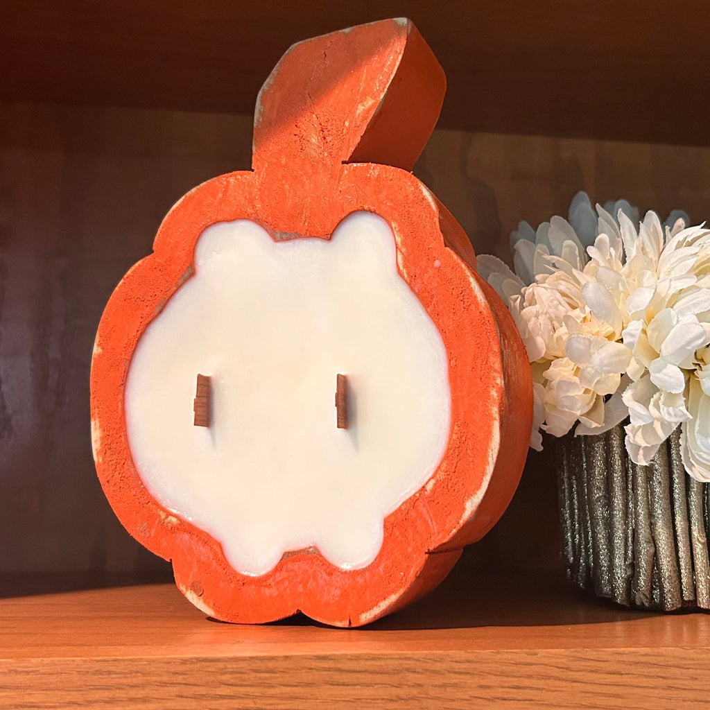 A pumpkin-shaped orange and white candle displayed on a wooden shelf next to a small vase of white flowers.