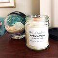 A glass jar candle labeled 'Chicwood Candle Co. Volcano Vibes' sits on a wooden table next to its matching decorative lid and a blue-green marbled glass orb, with a neutral indoor background.