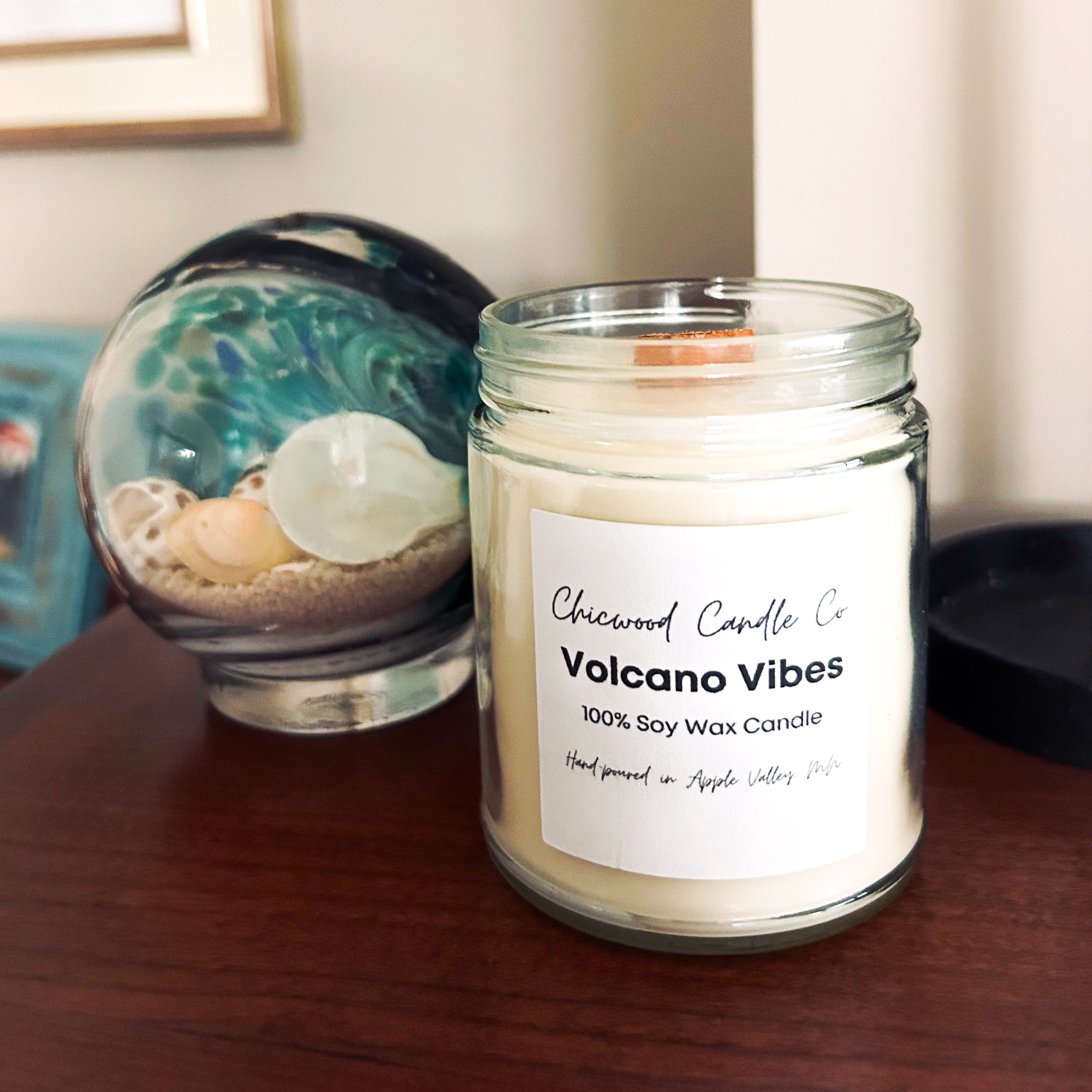 A glass jar candle labeled 'Chicwood Candle Co. Volcano Vibes' sits on a wooden table next to its matching decorative lid and a blue-green marbled glass orb, with a neutral indoor background.