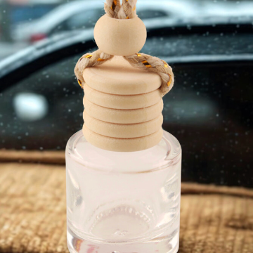 A clear glass car air freshener bottle with a beige wooden cap and a braided beige-and-gold hanging cord, resting on a textured surface with a blurred car window in the background.